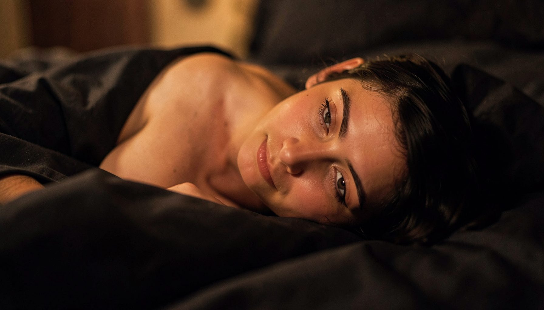 Warm intimate lighting, soft skin tones against dark sheets, suggestive romantic atmosphere